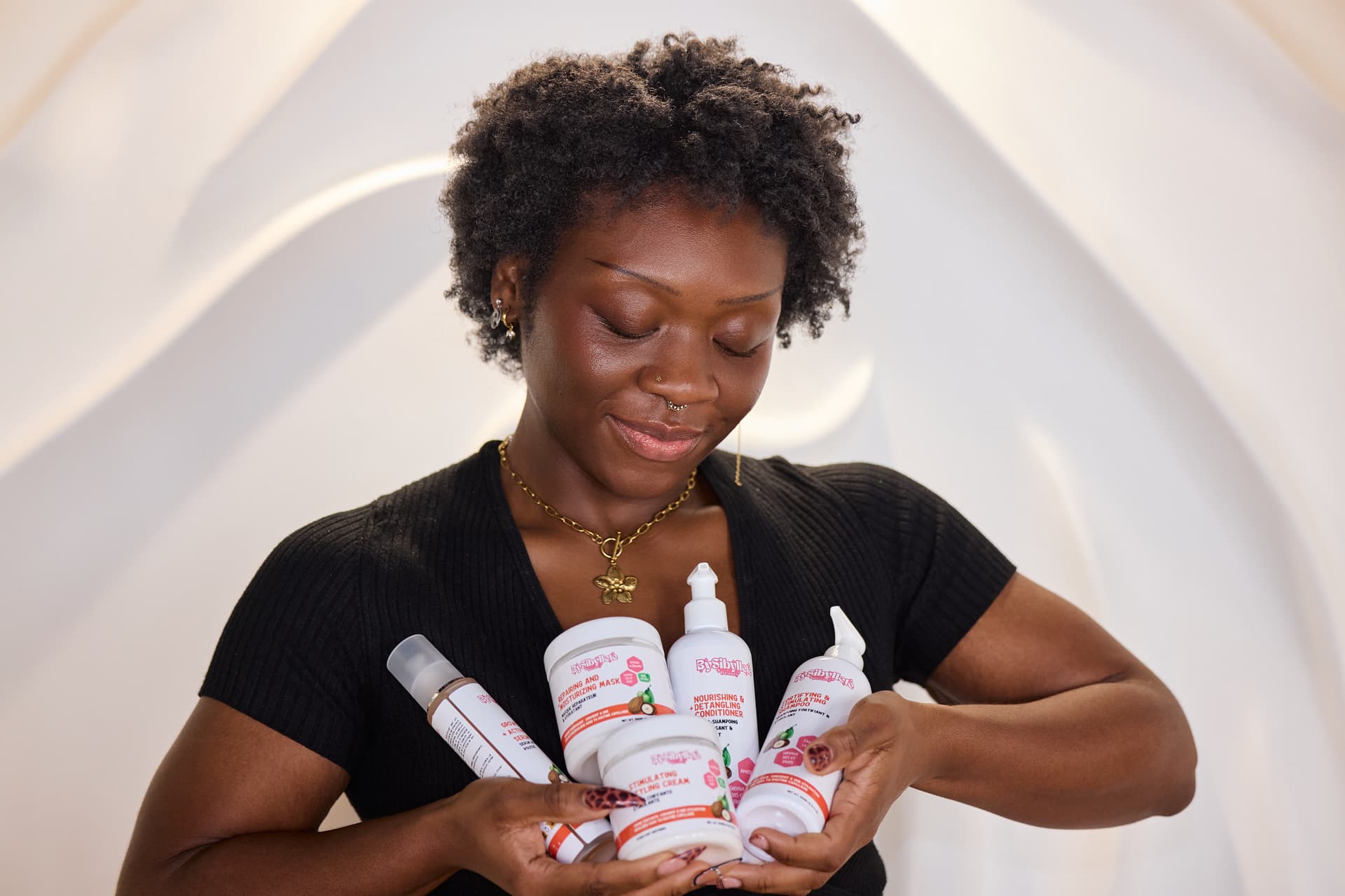 Une femme souriante tenant dans ses mains la gamme de soins capillaires By Sibyllete, comprenant le sérum, le masque, la crème coiffante et l'après-shampoing, pour une routine complète et naturelle.