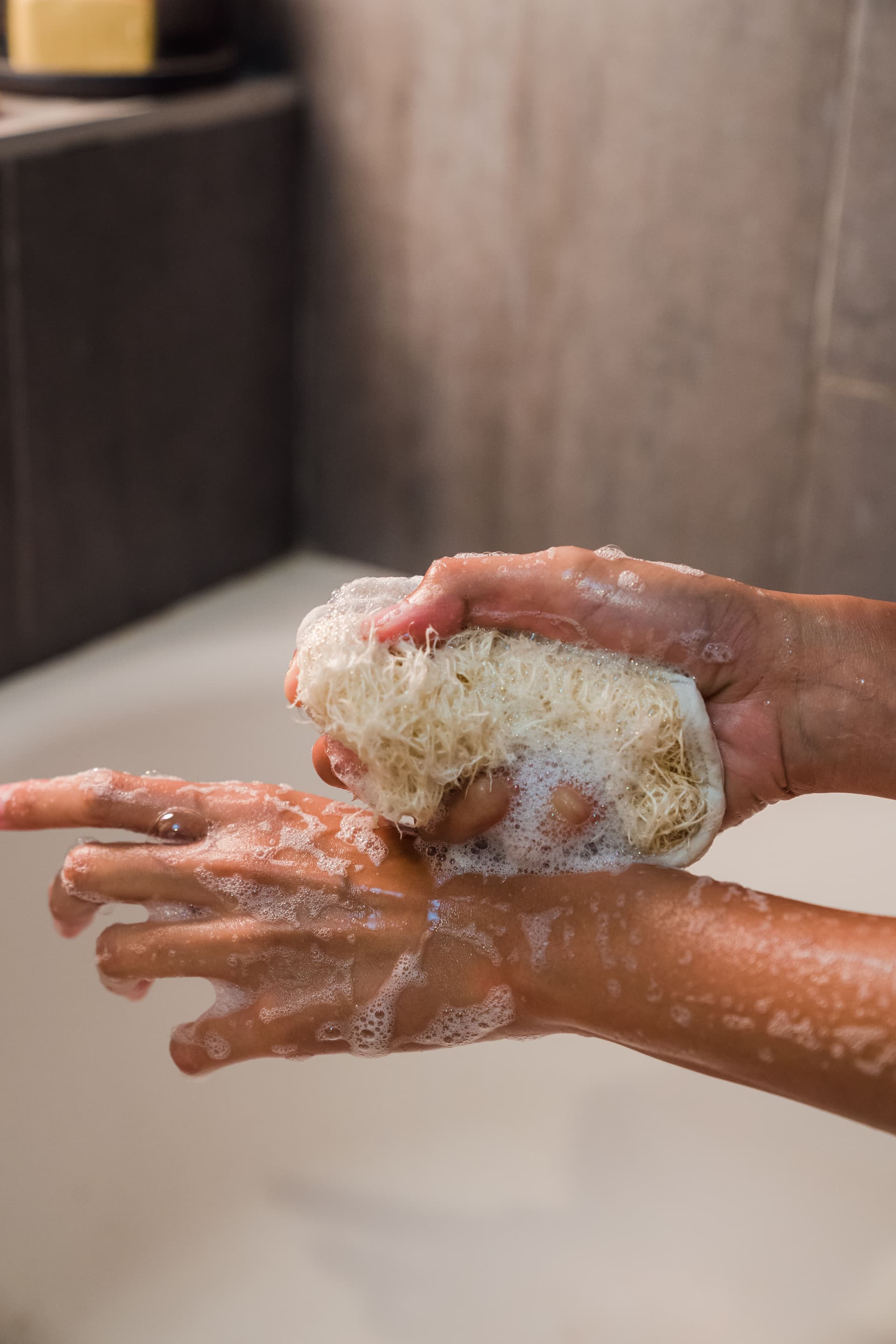 Prise de vue rapprochée de mains utilisant une éponge naturelle de luffa pour faire mousser un soin nettoyant et exfoliant sous la douche, pour un moment de bien-être.