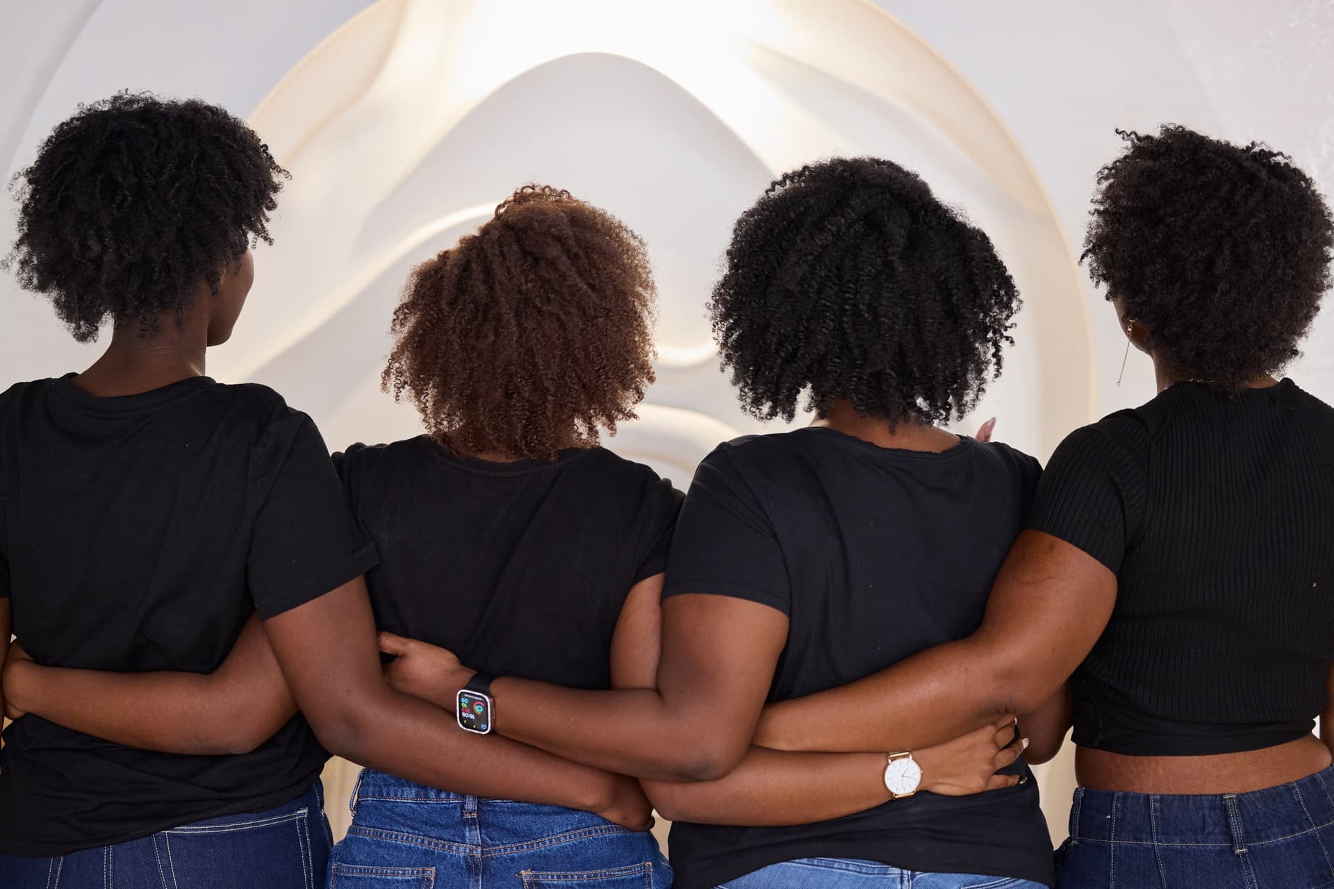 Quatre femmes vues de dos et enlacées, célébrant la beauté naturelle et la diversité des textures de cheveux afro et bouclés