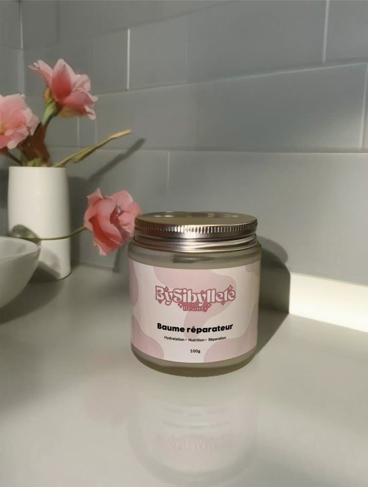 Pot du Baume réparateur By Sibyllete disposé dans un décor de salle de bain épuré avec des fleurs roses.