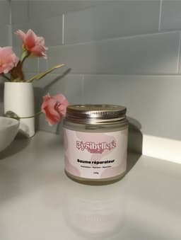Pot du Baume réparateur By Sibyllete disposé dans un décor de salle de bain épuré avec des fleurs roses.