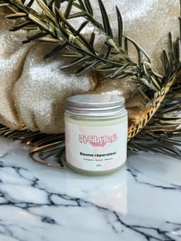 Baume réparateur 100g By Sibyllete posé sur du marbre, entouré de branches de romarin et de touches dorées.