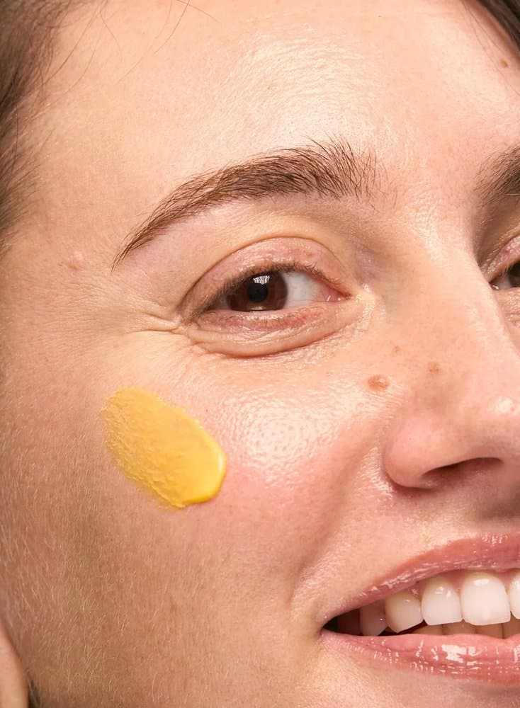 Gros plan sur deux touches de crème jaune au curcuma By Sibyllete appliquées sur la joue pour cibler l'éclat, l'unification et l'hydratation de la peau.