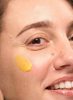 Gros plan sur deux touches de crème jaune au curcuma By Sibyllete appliquées sur la joue pour cibler l'éclat, l'unification et l'hydratation de la peau.