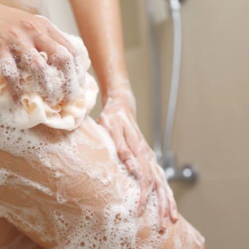 Gros plan sur une jambe généreusement savonnée à l'aide d'une éponge de douche, montrant une mousse riche et onctueuse.