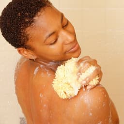 Une personne sous la douche utilisant une éponge naturelle pour faire mousser le produit de soin sur ses épaules, dans un moment de détente.