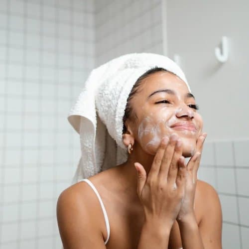 Personne souriante appliquant la mousse nettoyante sur le visage, une serviette sur la tête, dans un décor de salle de bain.