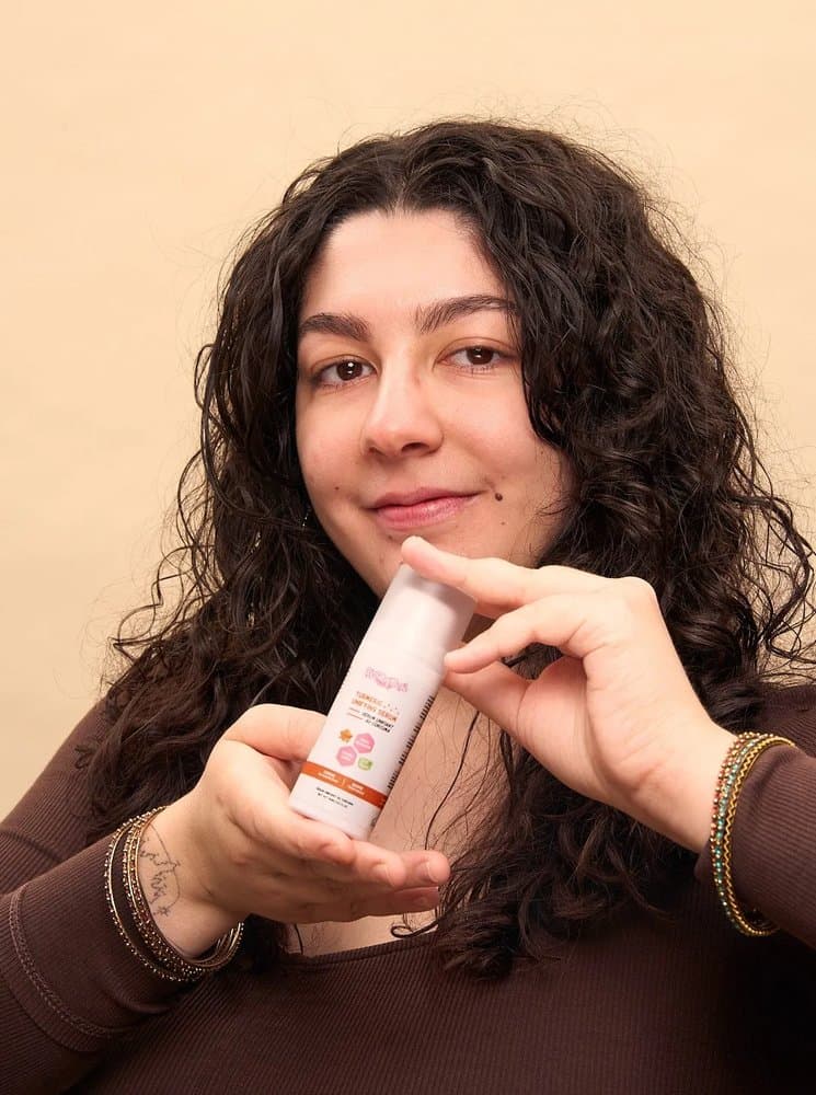Une personne souriante tenant le flacon de Sérum Unifiant au Curcuma (Turmeric Unifying Serum) By Sibyllete lors de sa routine beauté.